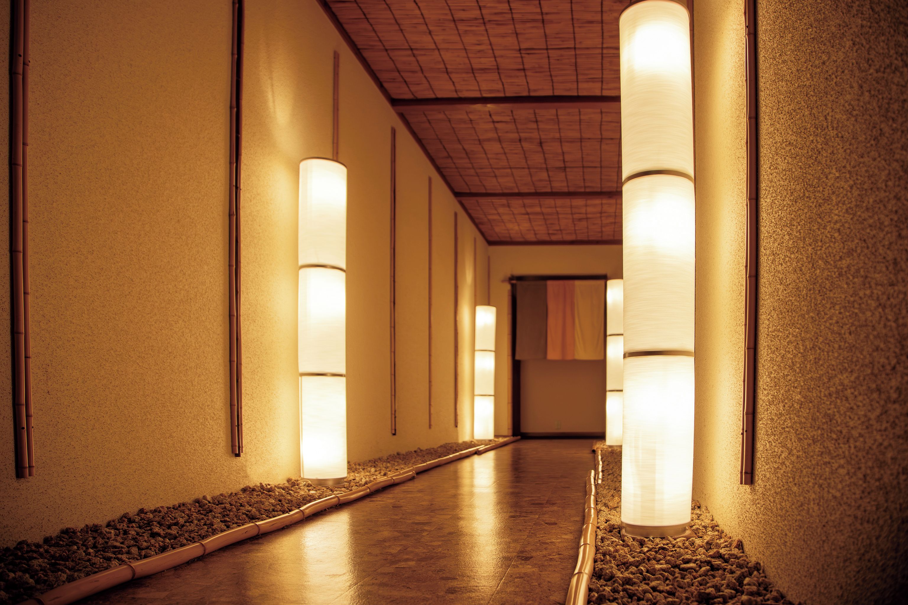 A corridor with a wooden floor, walls with textured surfaces, and cylindrical light fixtures along the walls.