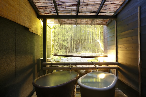 Un onsen japonés tradicional con dos tinas termales bajo un techo de bambú, rodeado de paredes de madera y vistas a un bosque de bambú.