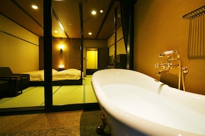 A hotel room with a large bathtub, a bed, a small table, and a mirror reflecting the room.