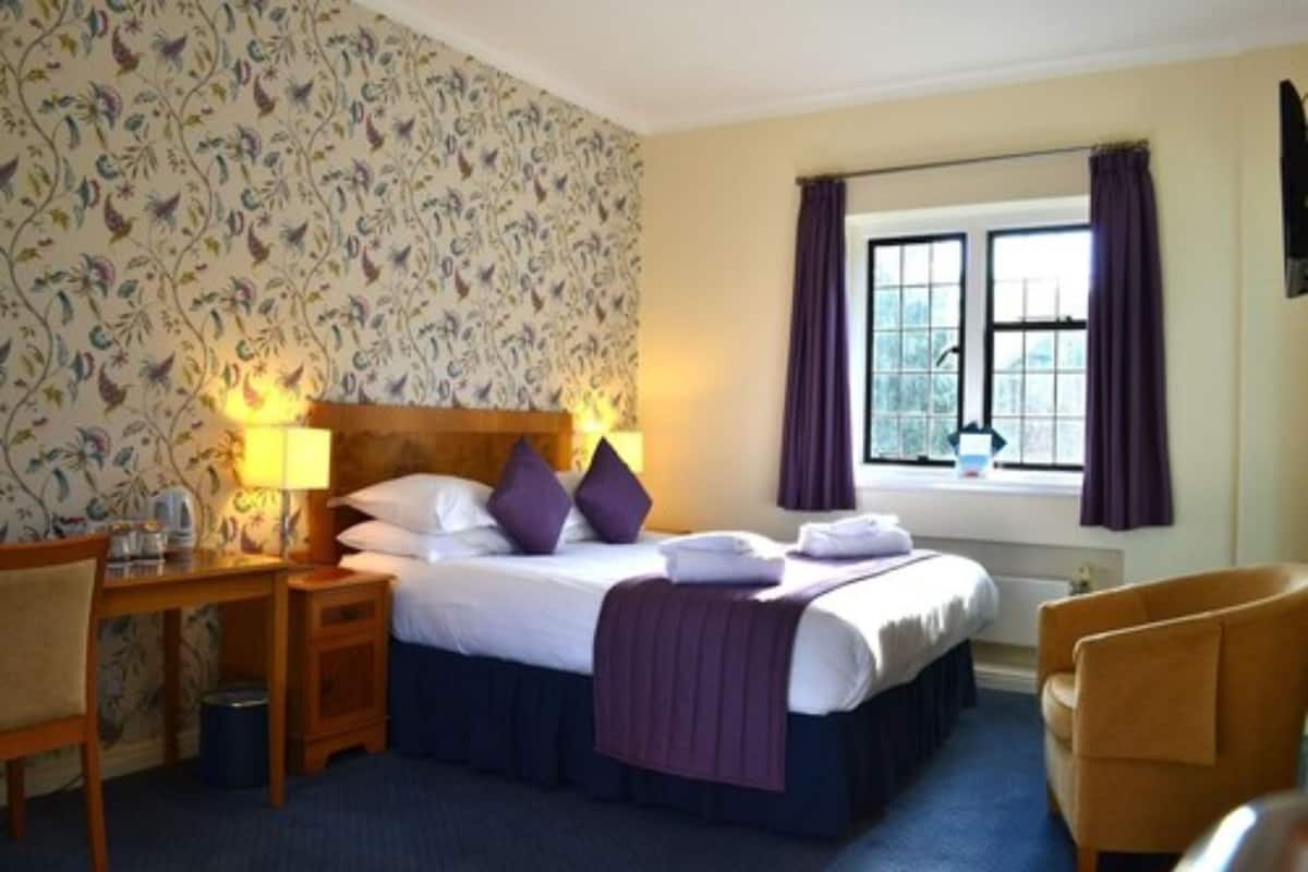 Una habitación de hotel con cama, mesitas de noche, una silla, una ventana con cortinas y un papel tapiz estampado.
