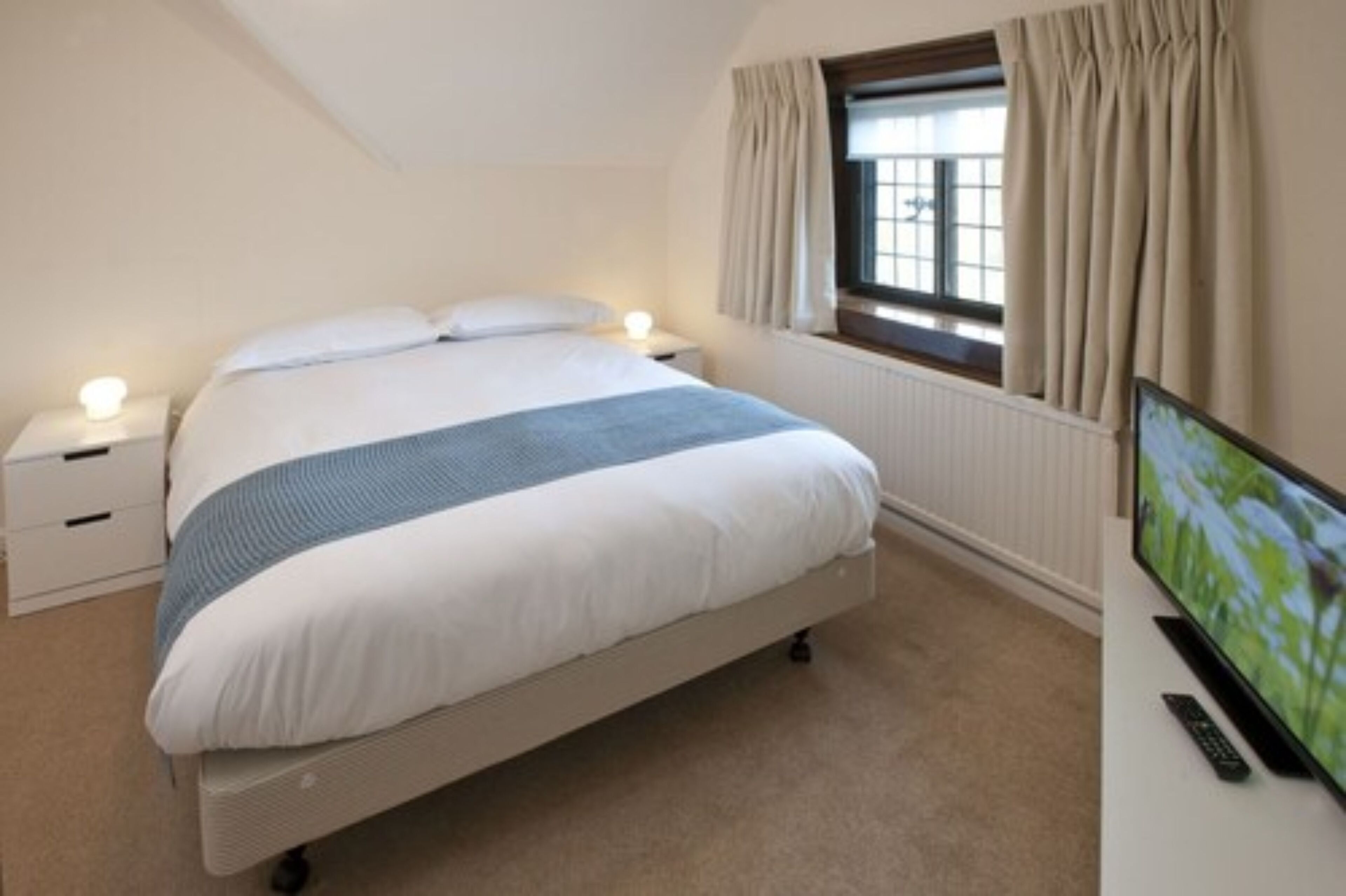 A bedroom with a bed, bedside tables, a TV, and a window with curtains.