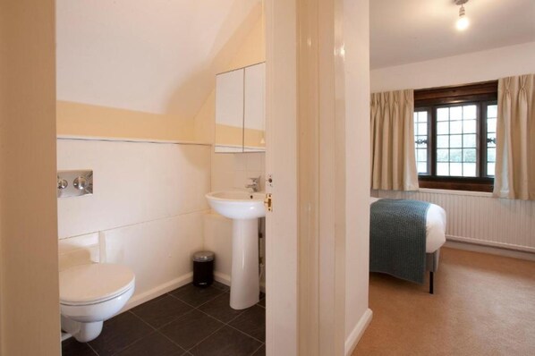 A small, clean bathroom with a toilet, sink, and mirror. There is a window with curtains and a radiator. - Madingley Hall (Cambridge)