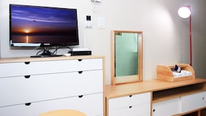 Una habitación moderna con una televisión sobre un escritorio de madera, un espejo, una lámpara de escritorio y una cama pequeña de madera.
