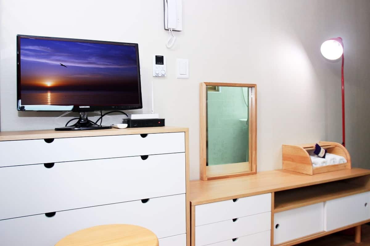 Ein modernes Zimmer mit Fernseher auf einem Holztisch, einem Spiegel, einer Schreibtischlampe und einem kleinen hölzernen Bett.
