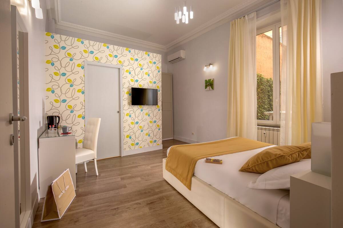 A bedroom with a bed, a TV mounted on the wall, a chair, and a desk with a coffee machine.