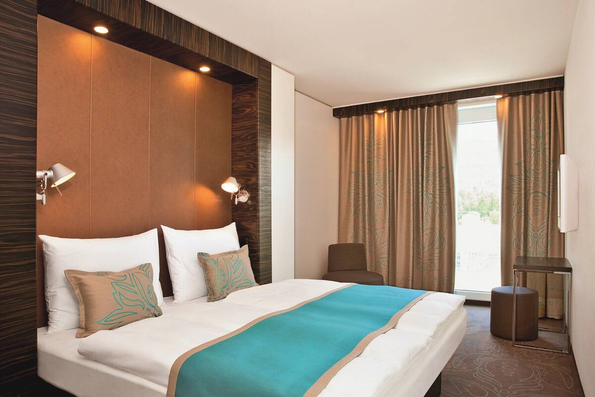 A modern bedroom with a large bed, a headboard with built-in lighting, a bedside table, and a view of the outside through curtains.