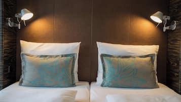 Ein Hotelzimmer mit zwei Betten, jedes mit einem weißen und einem dekorativen Kissen mit Blattform. An beiden Seiten der Betten befinden sich Wandleuchten.