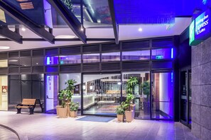 Una entrada de hotel moderna con fachada de vidrio, asientos al aire libre y un área de restaurante visible en el interior.