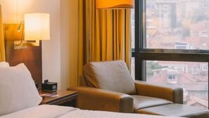A hotel room with a bed, a chair, a nightstand, a lamp, and a view of a cityscape through a large window. - Holiday Inn Express & Suites Bogota DC (Bogotá)
