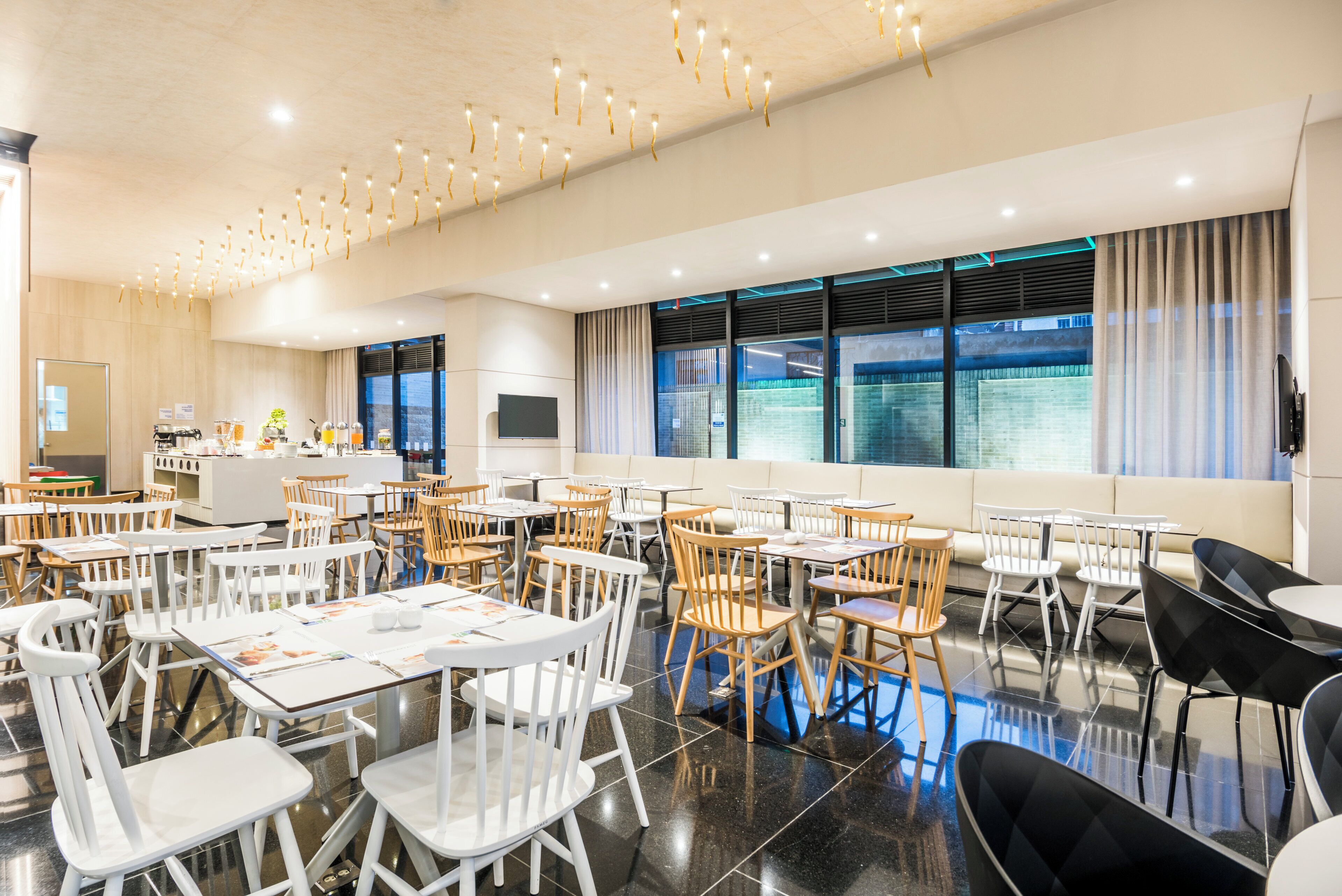 A spacious dining area with multiple tables and chairs, a kitchen area, and large windows with curtains.
