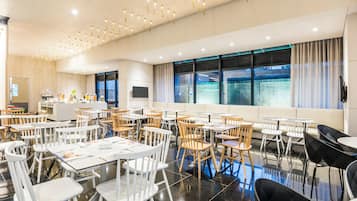 A spacious dining area with multiple tables and chairs, a kitchen area, and large windows with curtains.