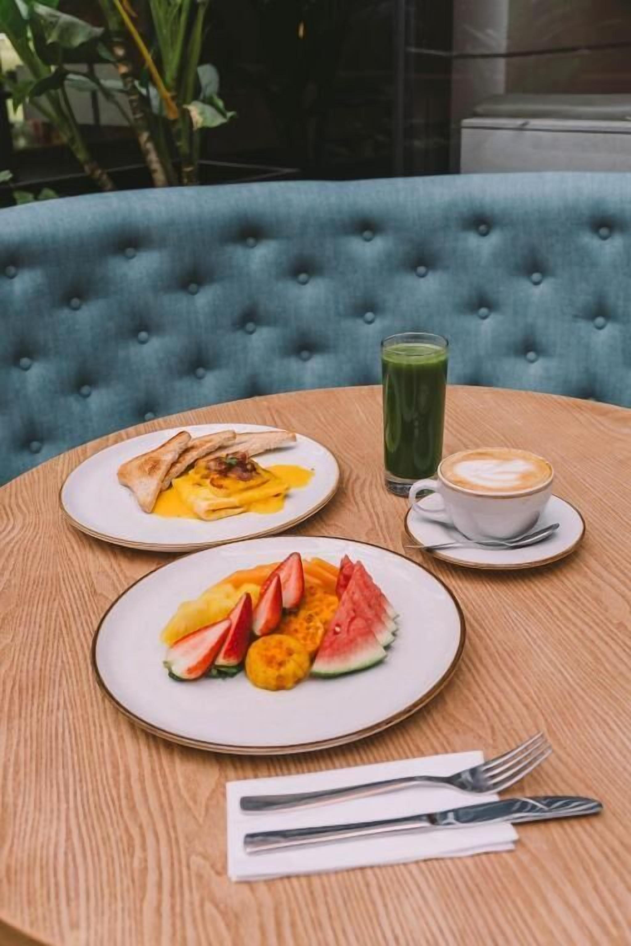 A round wooden table with a blue upholstered chair, two plates of breakfast food, a glass of green smoothie, and a cup of coffee.