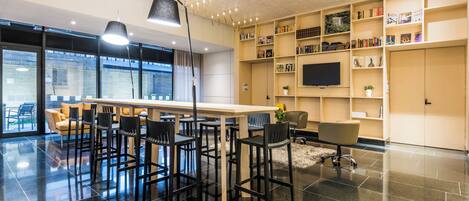A modern library with a long table, multiple chairs, bookshelves, a flat-screen TV, and large windows.