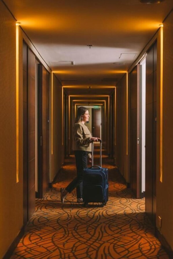 Un pasillo de hotel con alfombra estampada, una persona con maleta y una puerta al final.