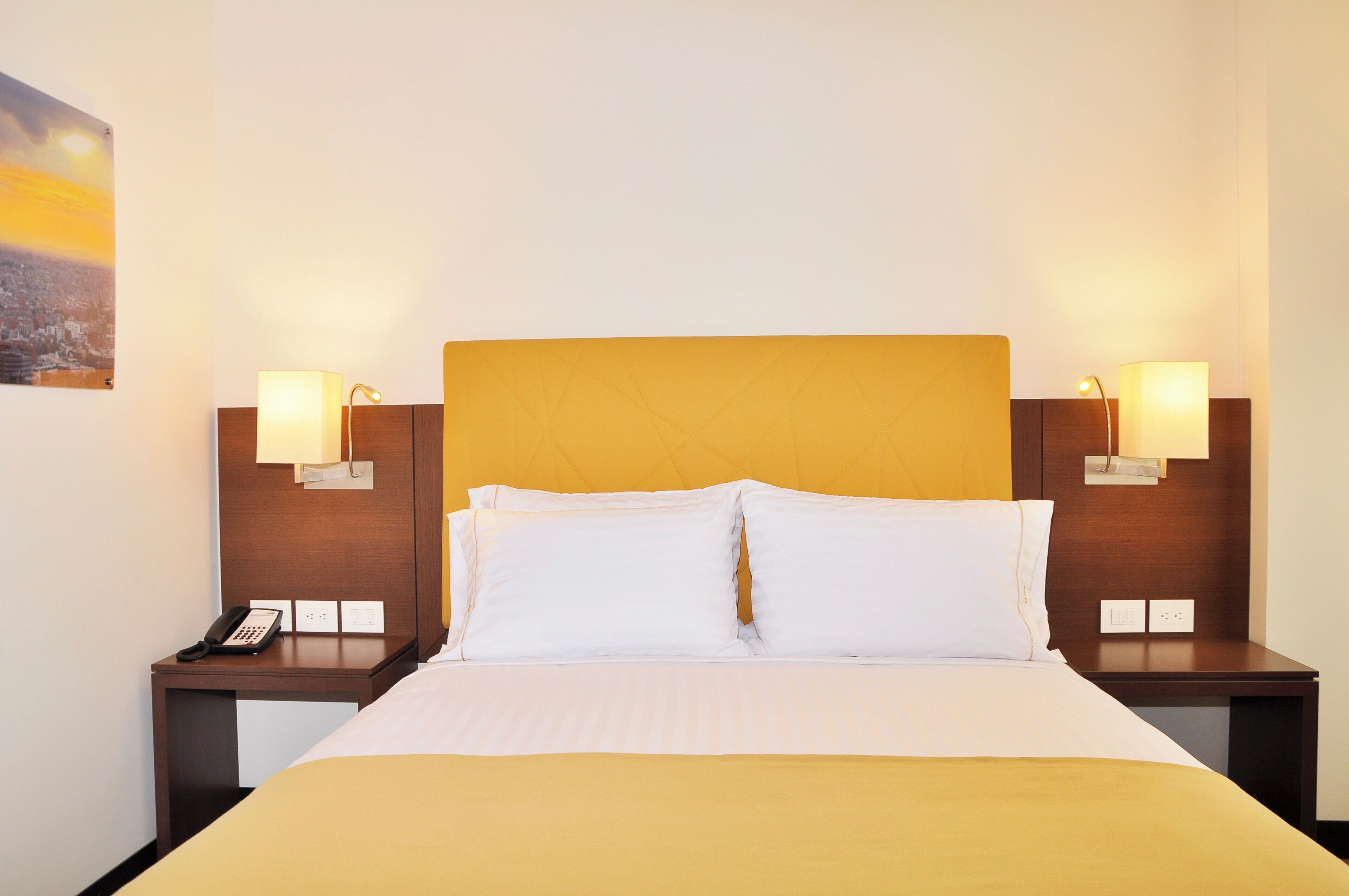 Quarto de hotel com cama, duas mesinhas de cabeceira, telefone e duas luminárias de parede.