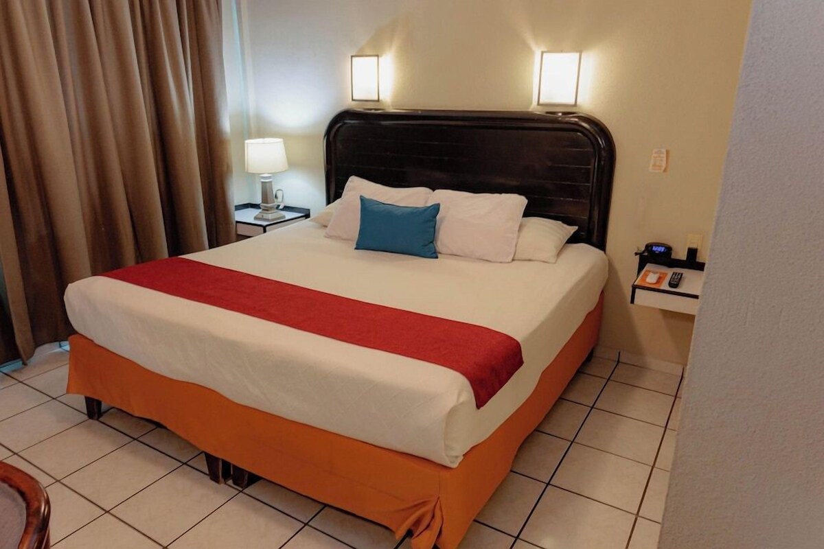 A hotel room with a bed, a nightstand, a lamp, and a wall-mounted light fixture.