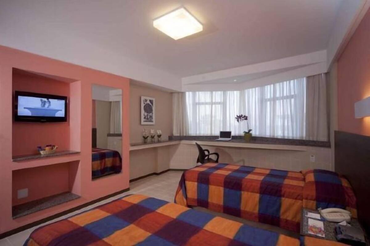 Une chambre d’hôtel avec deux lits, une télévision, un bureau avec un ordinateur portable, une fenêtre avec des rideaux et un miroir.