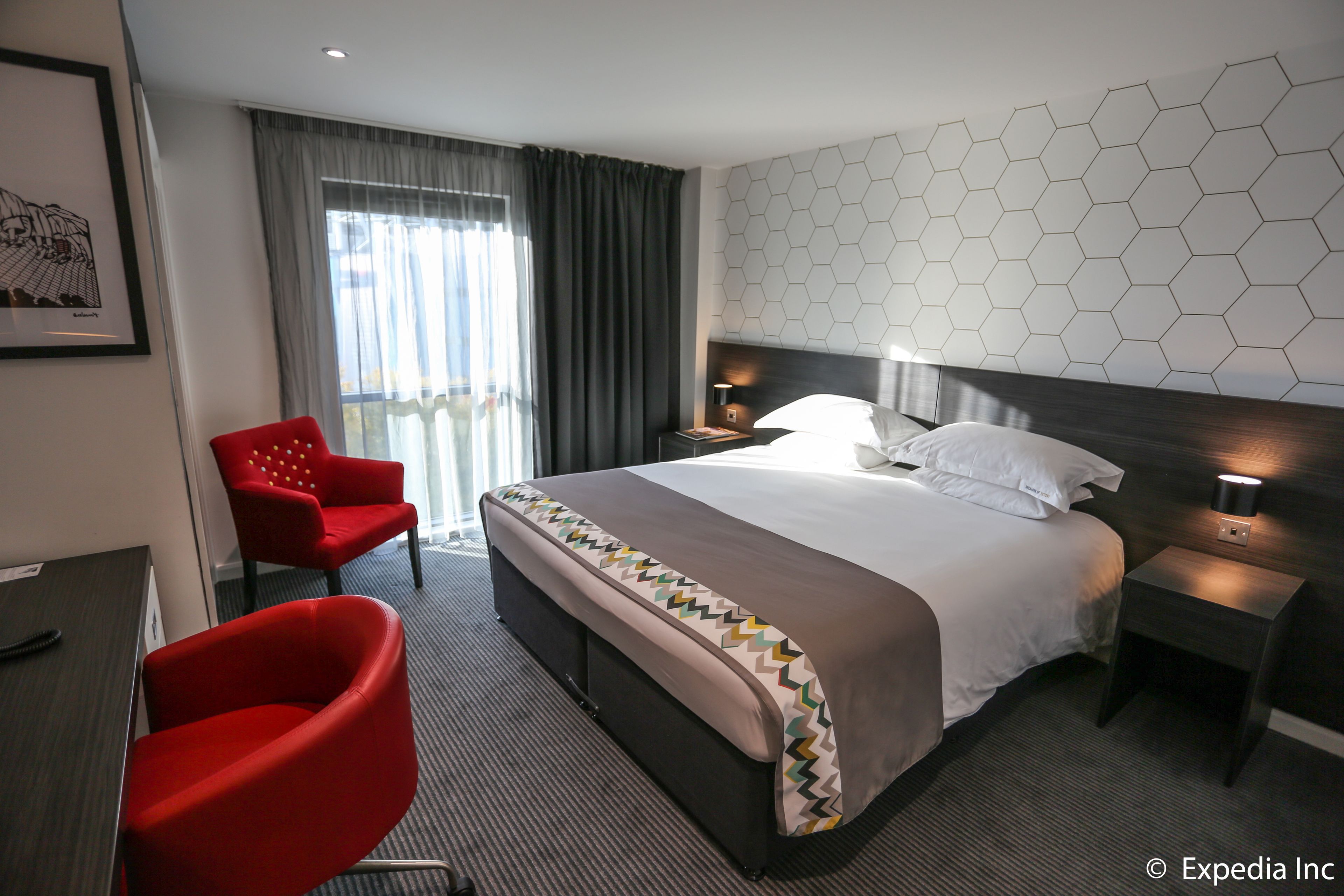 A hotel room with a large bed, a red armchair, a desk, and a view of the outdoors through a window with curtains.