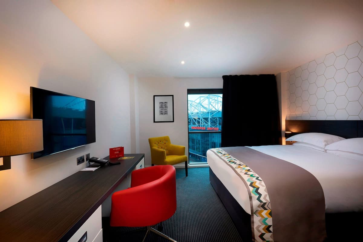 A hotel room with a bed, a desk with a TV, a red chair, and a window with a view of a building.