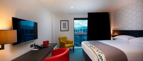 Habitación de hotel con una cama, un escritorio con televisor, una silla roja y una ventana con vistas a un edificio.