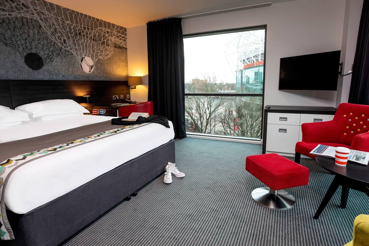 A hotel room with a large bed, a red armchair, a small table with a cup and newspaper, and a TV on the wall.