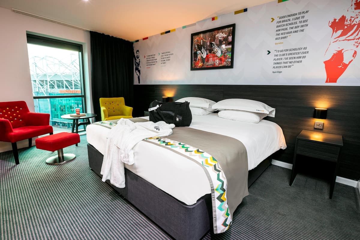 A hotel room with a bed, a red armchair, a yellow armchair, a small table, and a window with a view of a building.