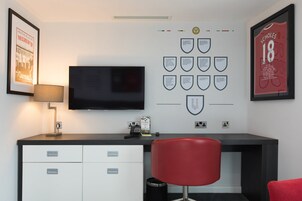 Una habitación moderna con televisor de pantalla plana, escritorio y una silla roja. En la pared hay elementos enmarcados, incluyendo una camiseta deportiva y un cartel.