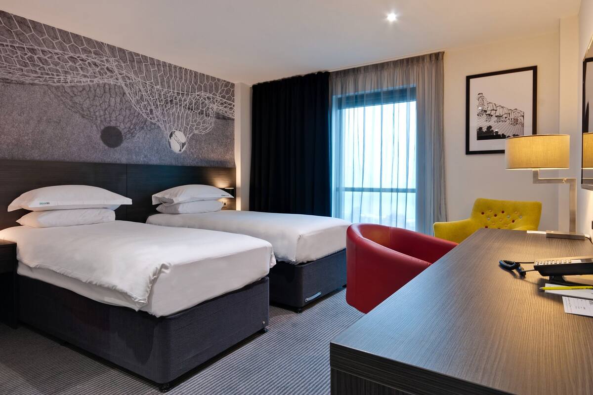 A hotel room with two beds, a desk, a chair, and a large window with curtains.