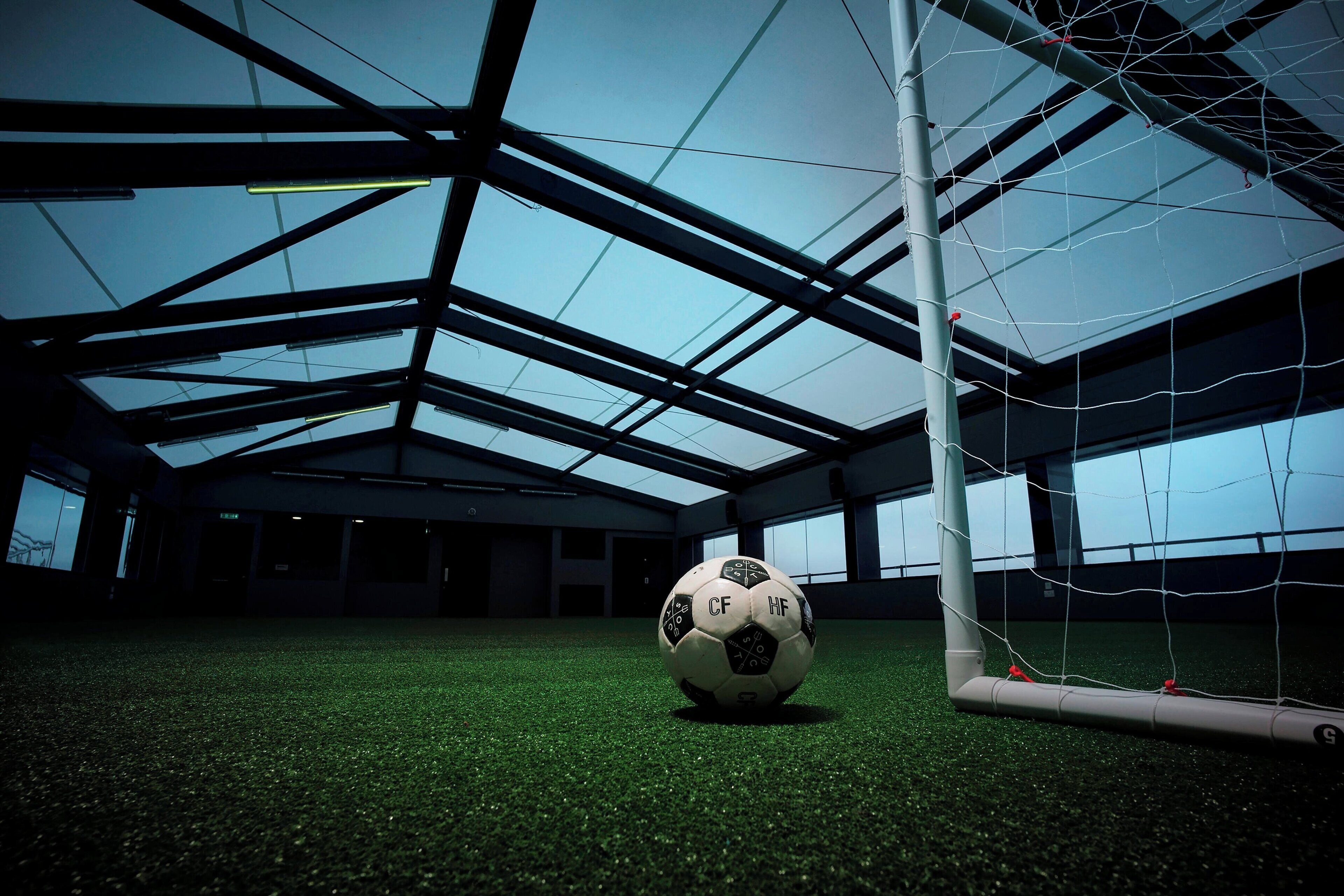 Un ballon de football devant un filet de but dans une installation intérieure dotée d’un plafond haut et d’une pelouse artificielle.