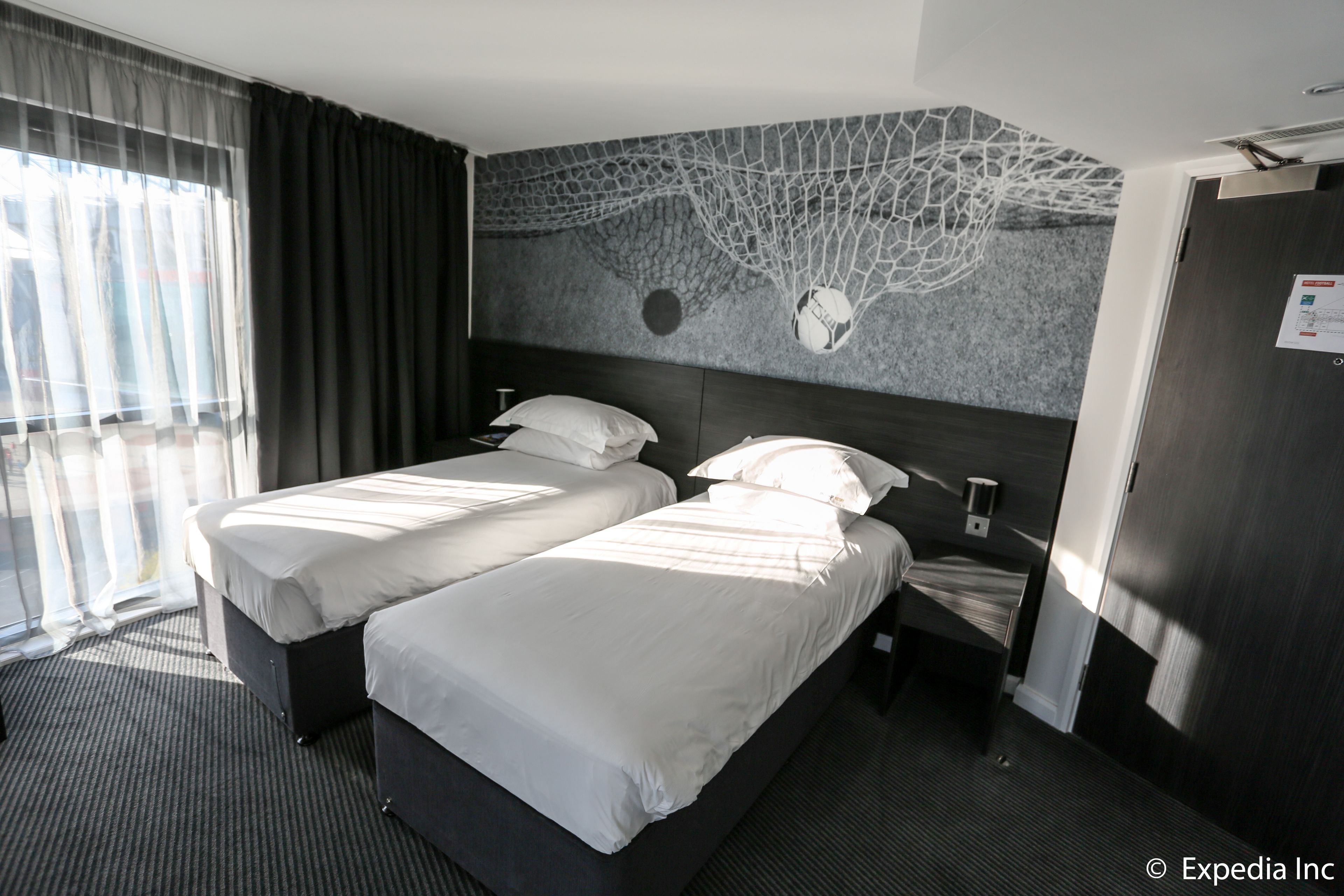 Habitación de hotel con dos camas, un ventanal con cortinas, un mural en la pared y una puerta con un letrero.