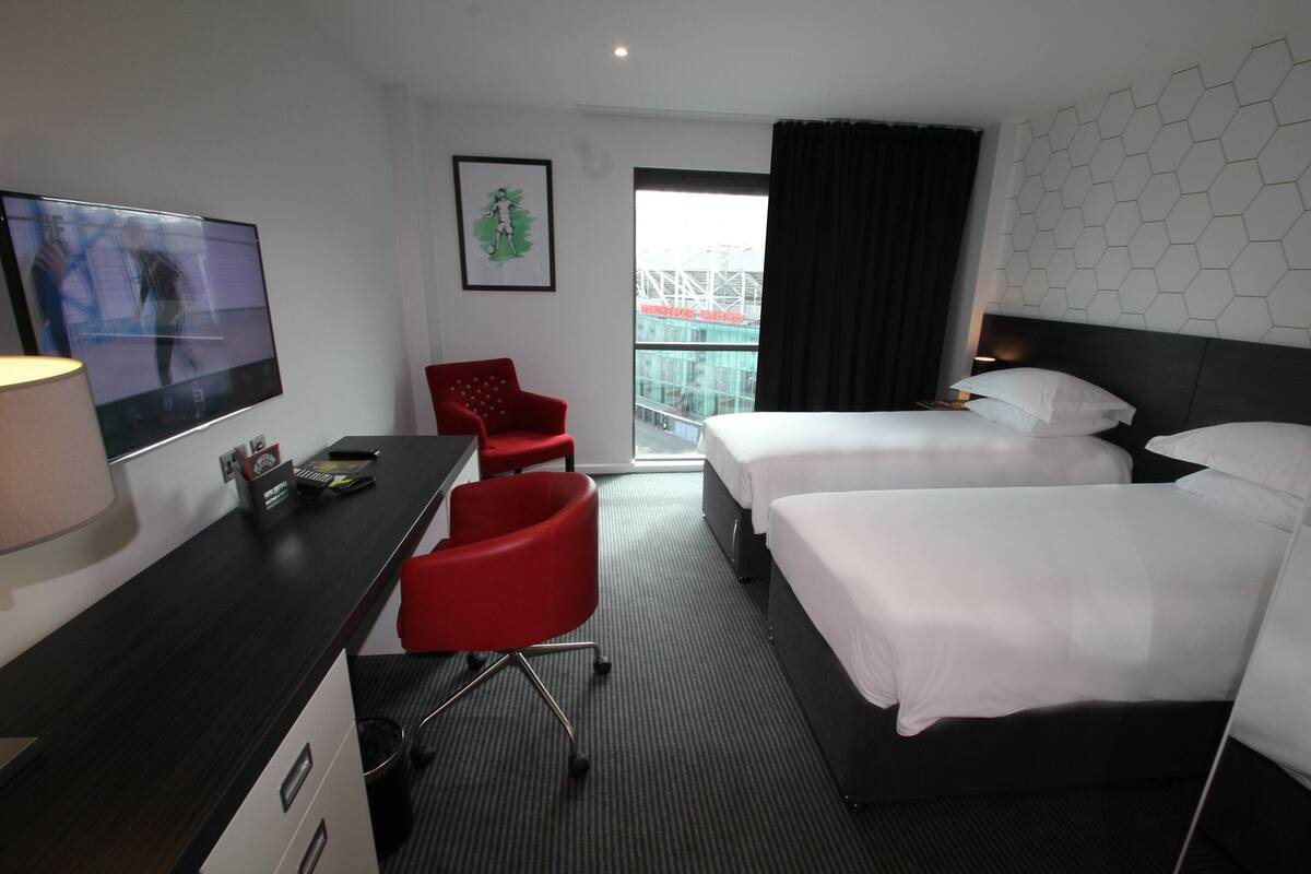 A hotel room with a desk, two beds, a red chair, a TV, and a window with a view of a building.