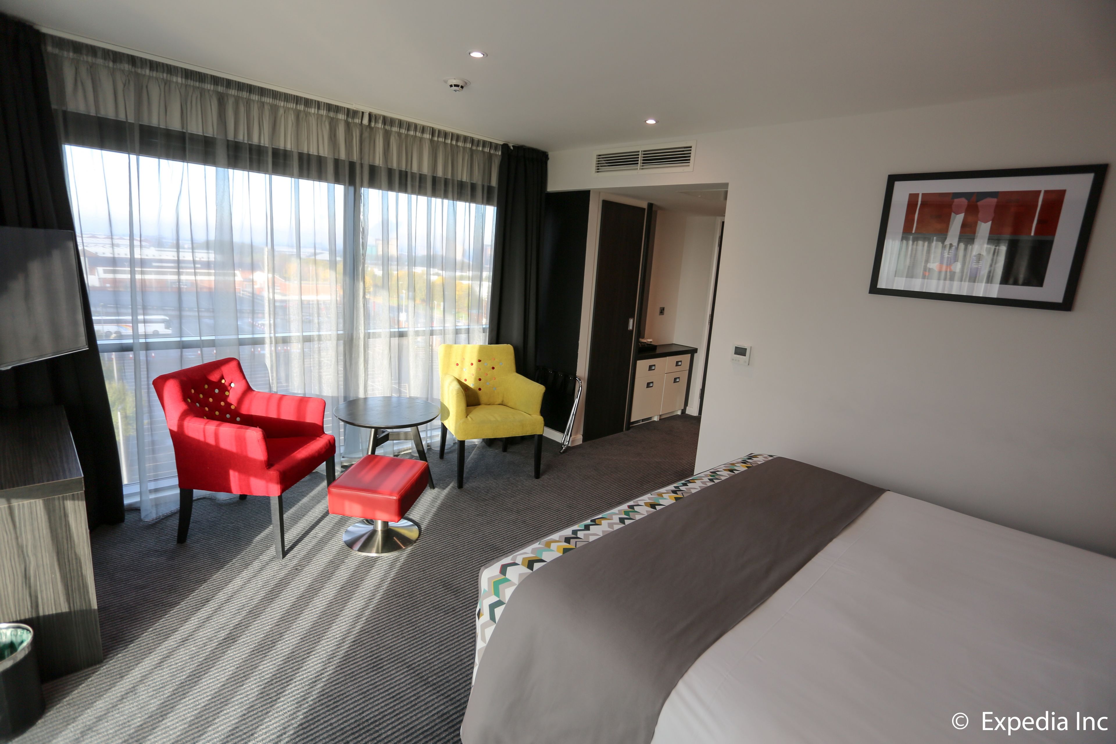 A hotel room with a large bed, a red armchair, a yellow armchair, a small table, and a view of the airport through the window.
