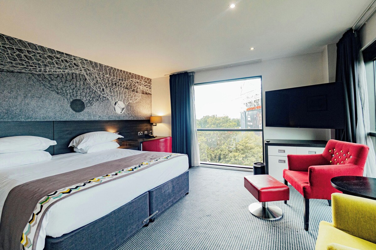A hotel room with a bed, a desk, a red chair, a TV, and a window with a view of trees.