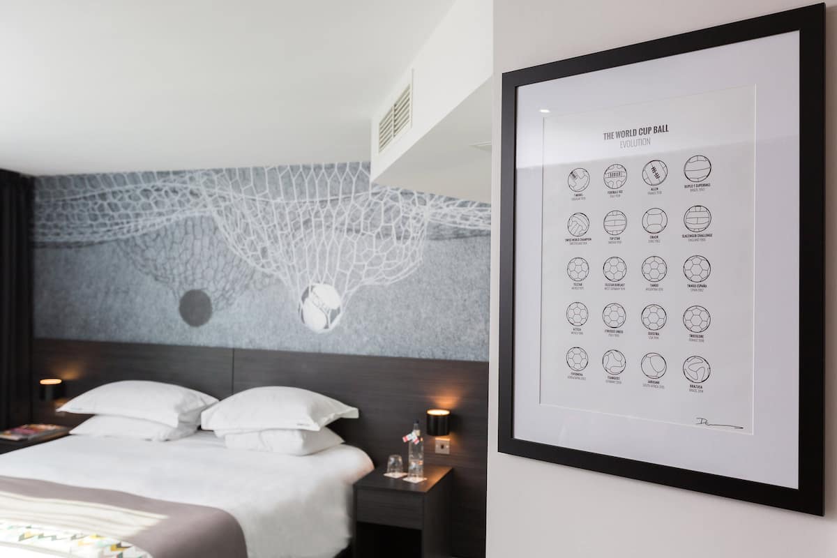 A hotel room with a bed, nightstands, a framed world cup evolution poster, and a wall with a textured design.