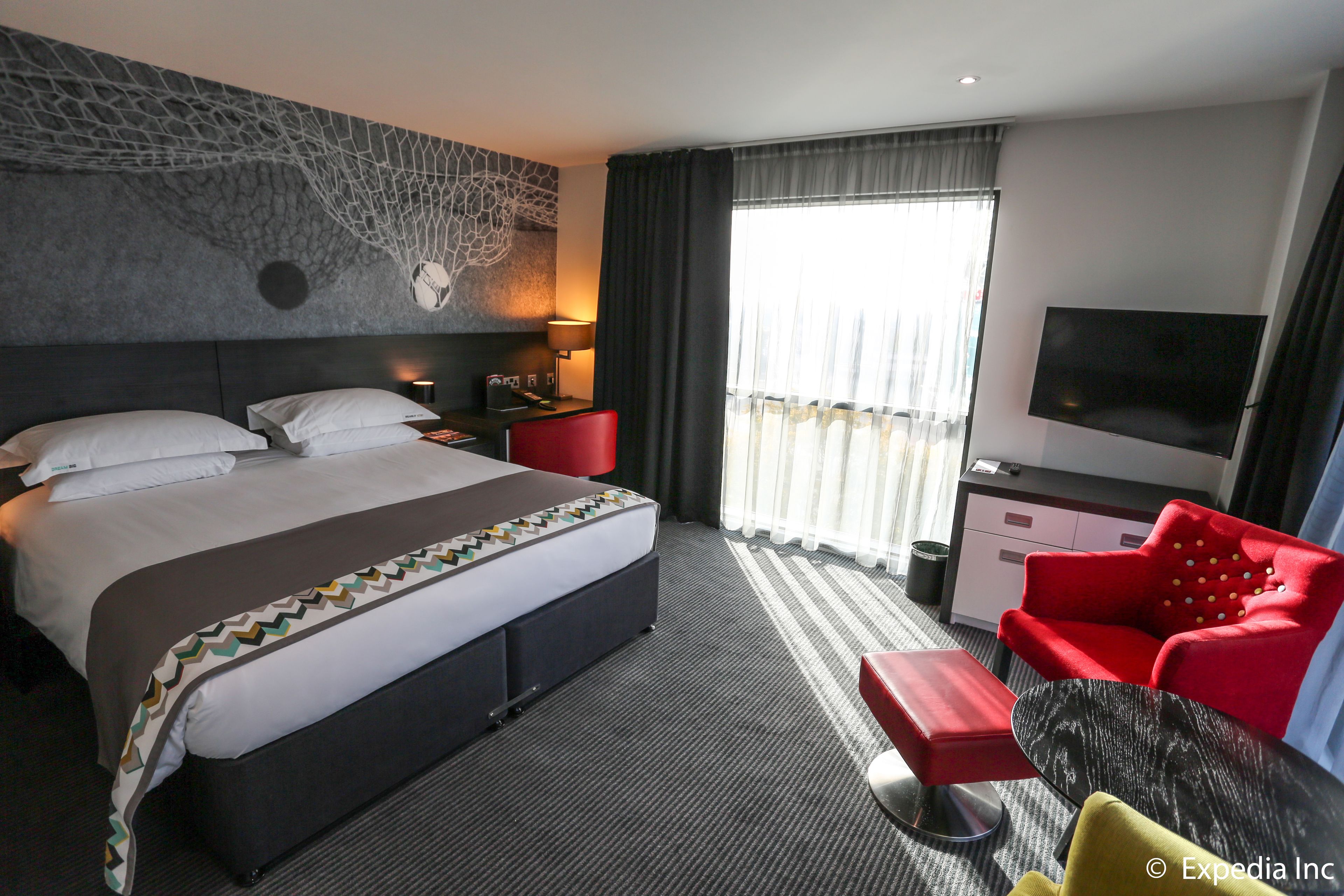 Habitación de hotel con una cama grande, un sillón rojo, un televisor y una ventana con cortinas.