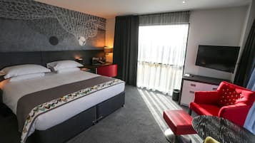 Habitación de hotel con una cama grande, un sillón rojo, un televisor y una ventana con cortinas.