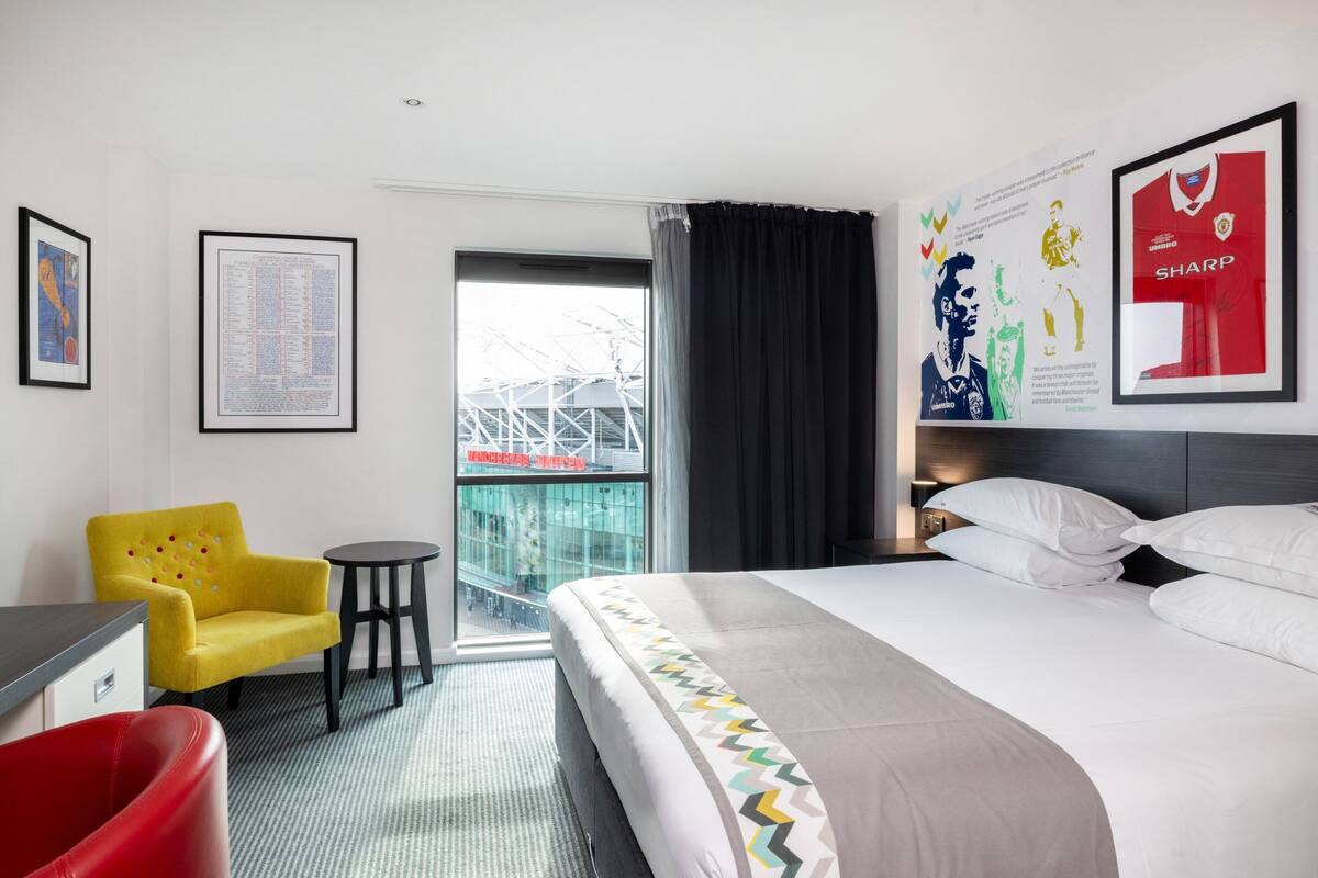 A hotel room with a bed, a yellow armchair, a red chair, a small table, and a framed poster on the wall.