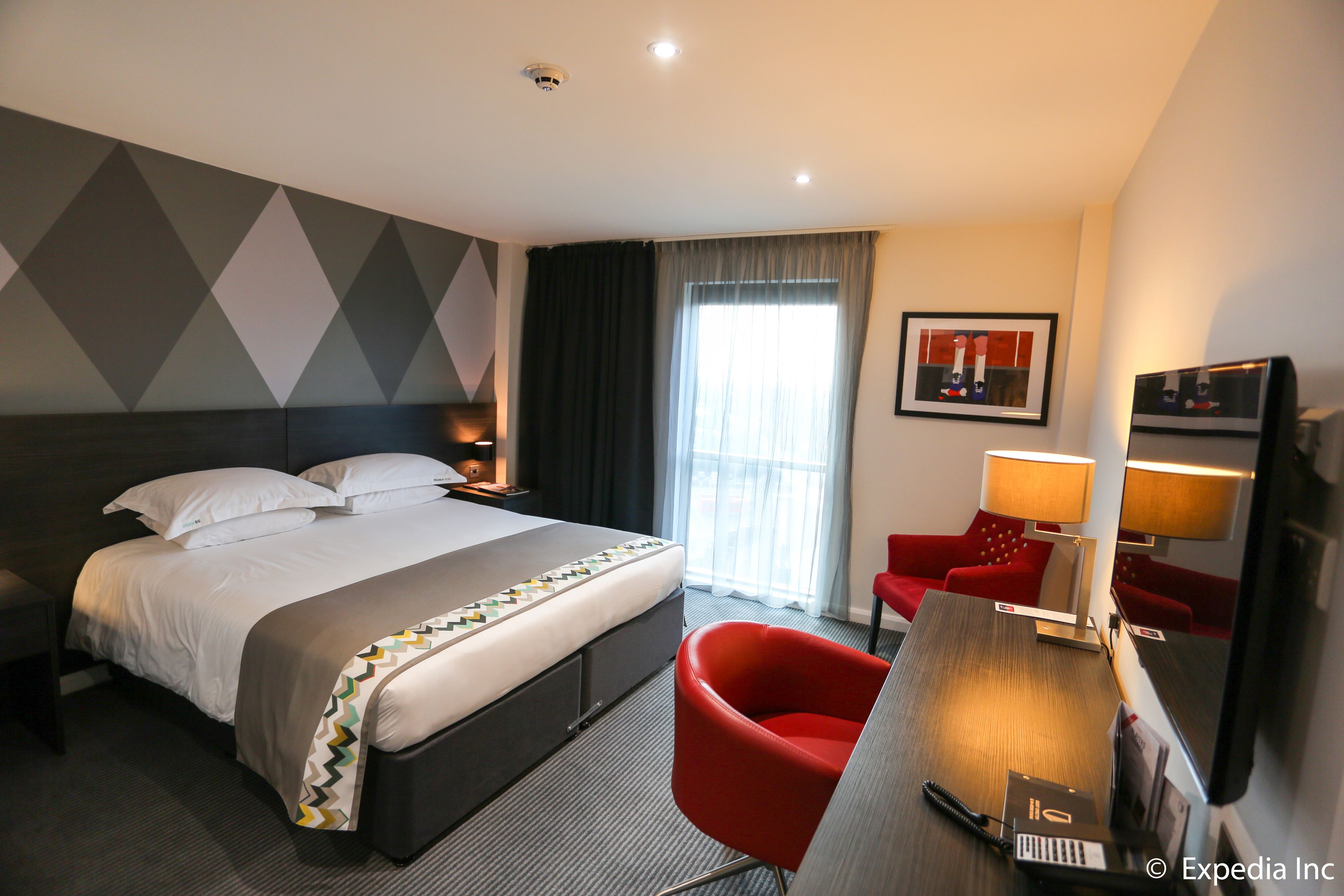 A modern hotel room with a large bed, a desk with a chair, a desk lamp, and a television.