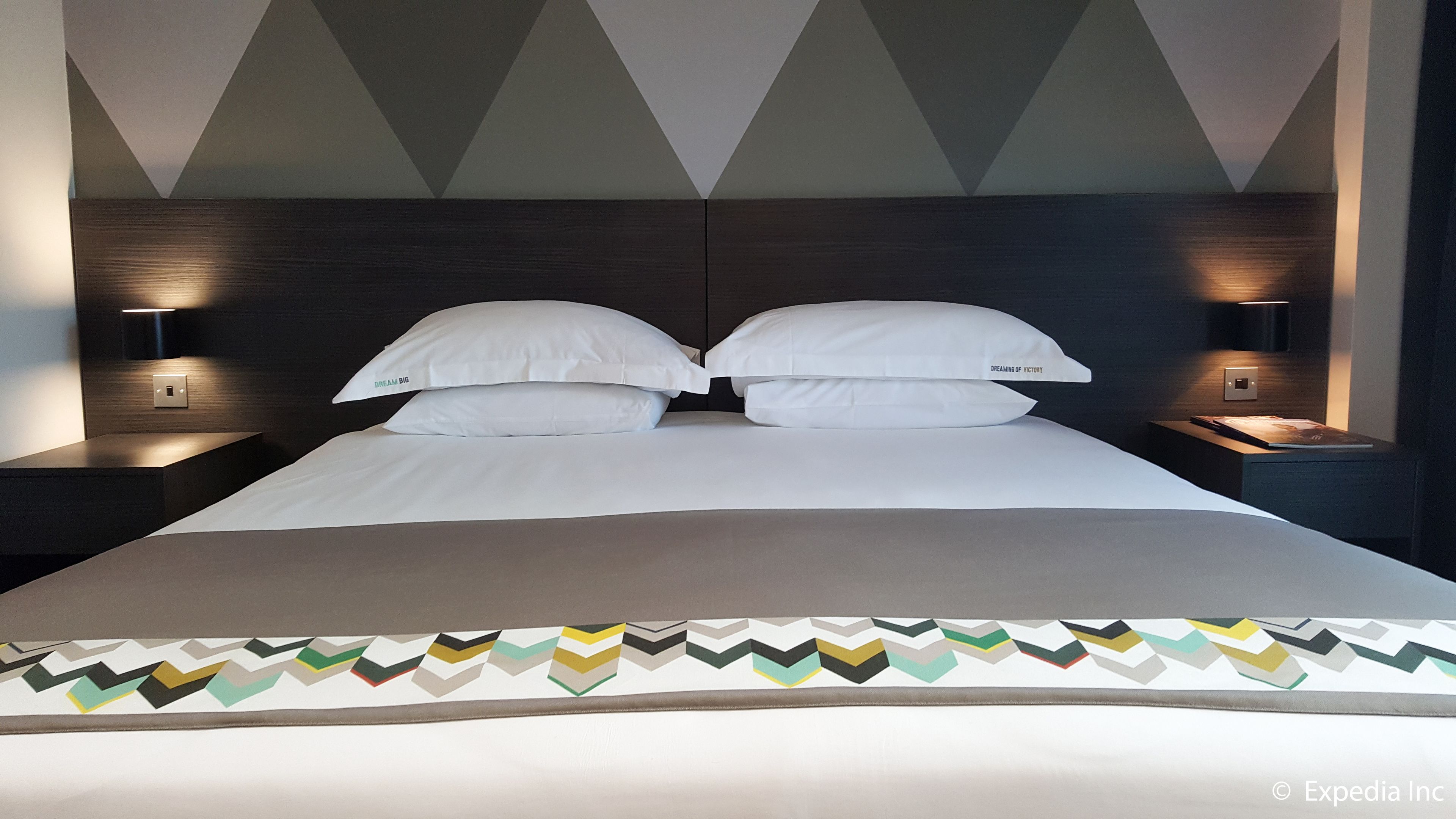 Un lit double bien fait, avec du linge de lit blanc, deux tables de chevet avec des lampes et un couvre-lit à motifs géométriques.
