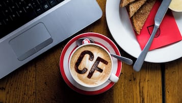 Una computadora portátil, una taza de café con arte de latte, una tostada y un cuchillo para mantequilla sobre una mesa de madera.