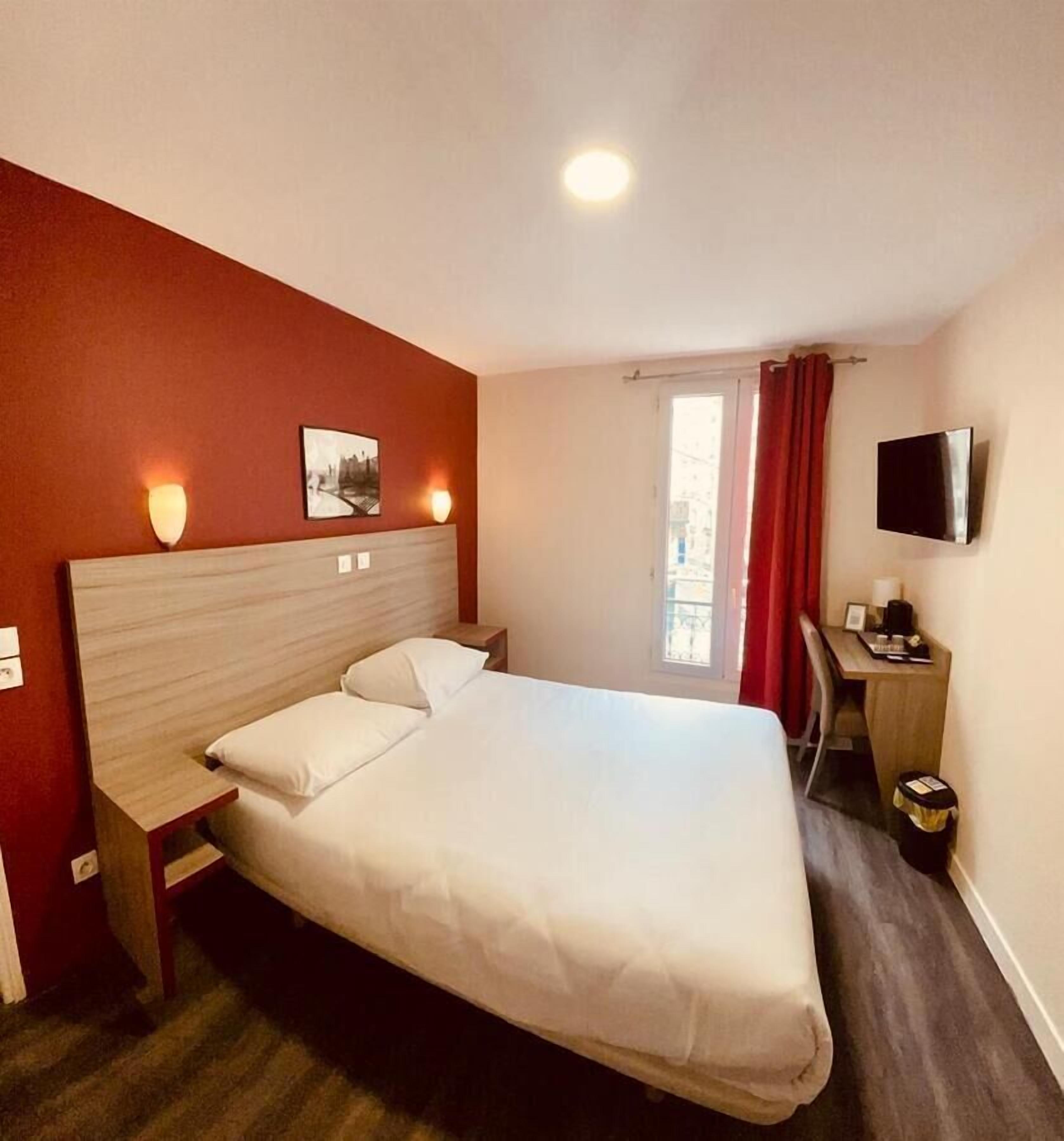 Habitación de hotel con una cama grande, un escritorio con silla, un televisor montado en la pared y un ventanal con cortinas rojas.