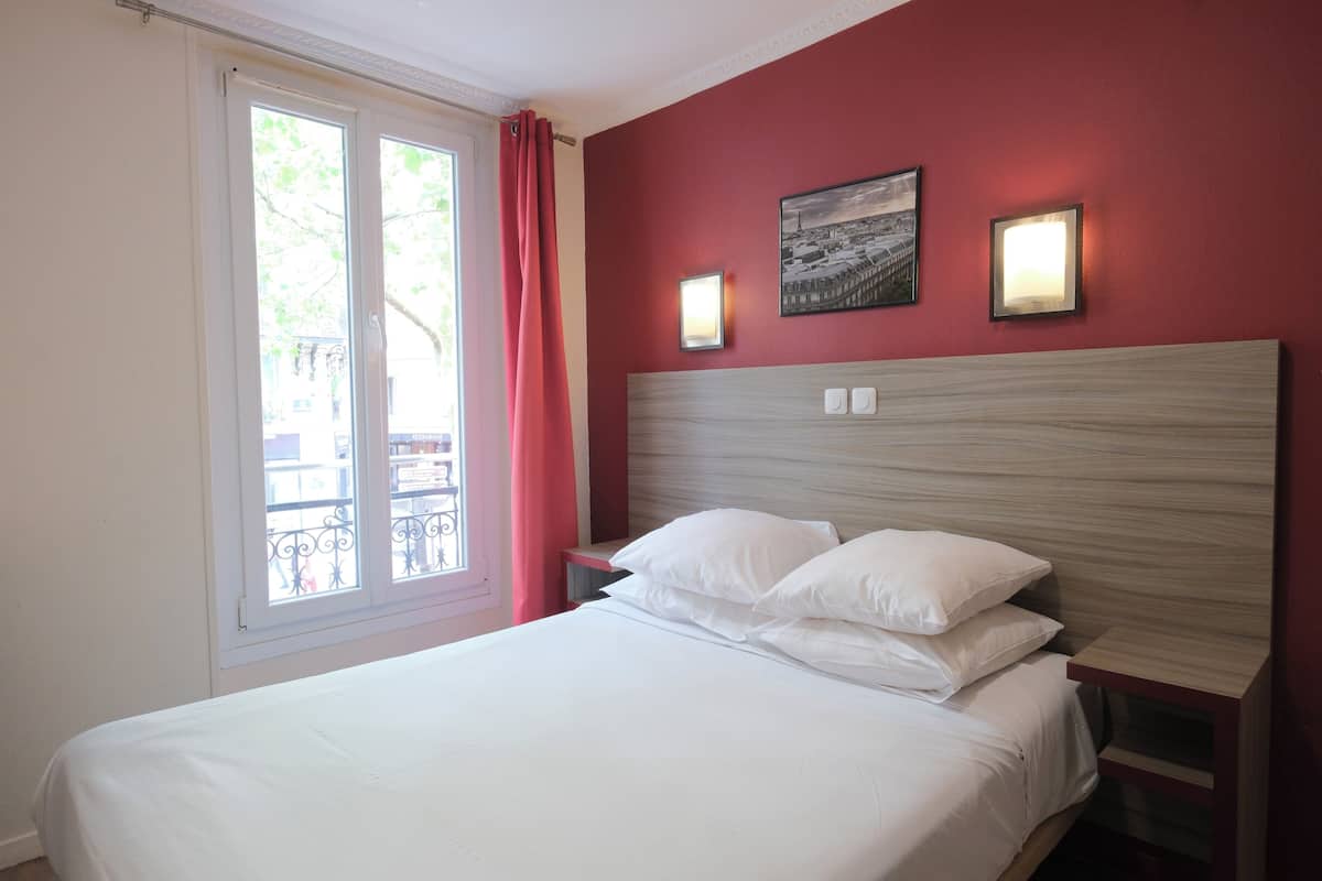 A hotel room with a bed, a window with curtains, a nightstand, and two wall-mounted lights.