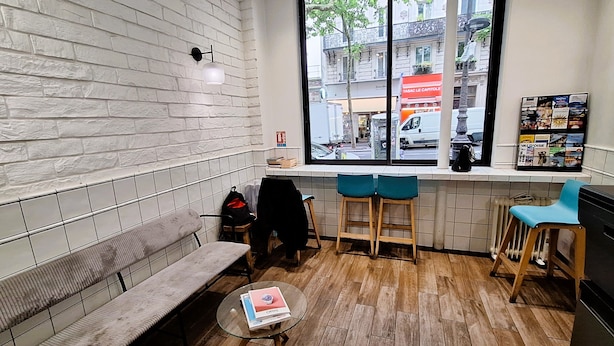 A cozy cafe with a white brick wall, a wooden bench, a small round table with a book, and a window overlooking a street with a shop. - Hipotel Paris Gambetta Republique (Paris)