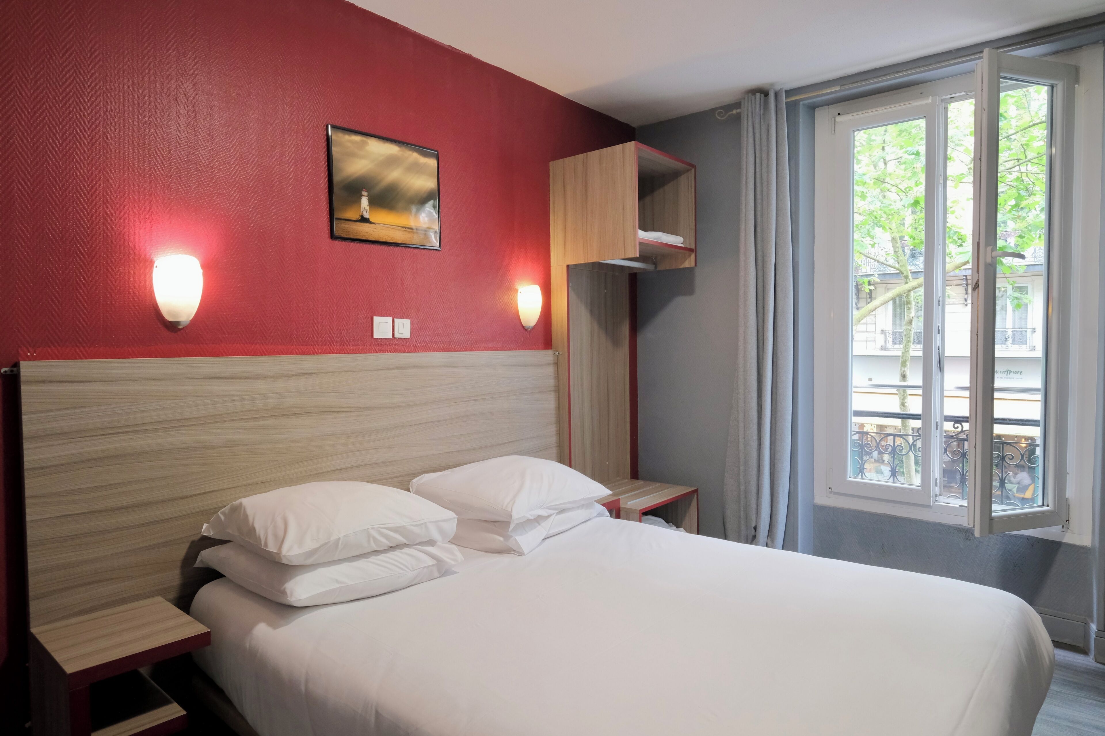 A hotel room with a bed, a nightstand, a wall-mounted light, a window with a view, and a painting on the wall.