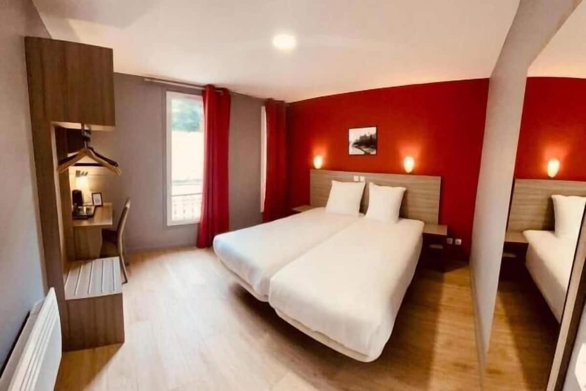 A hotel room with a bed, bedside tables, a desk with a chair, a wardrobe, and a window with curtains.
