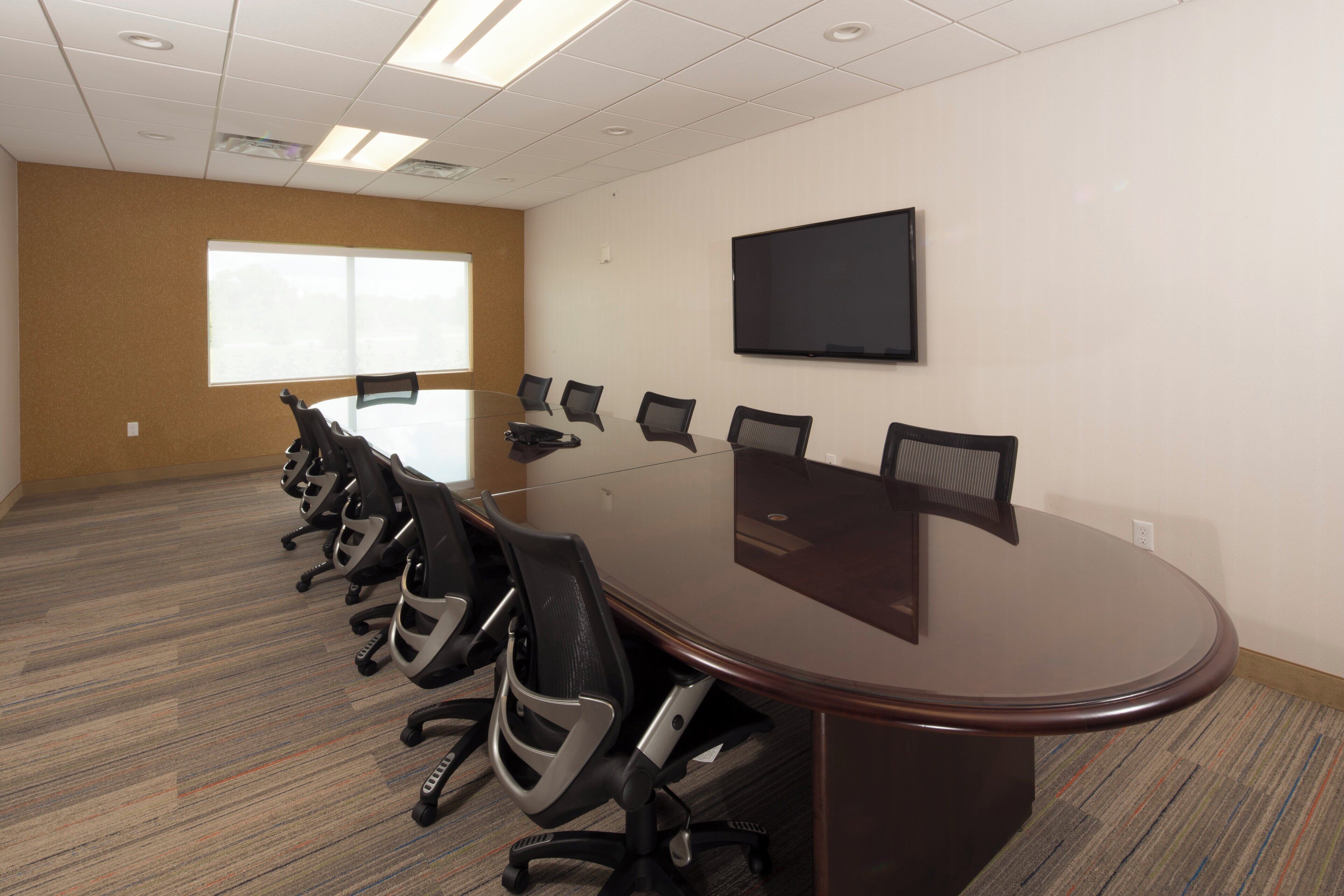 A conference room with a long table, multiple chairs, a flat-screen TV, and a large window.