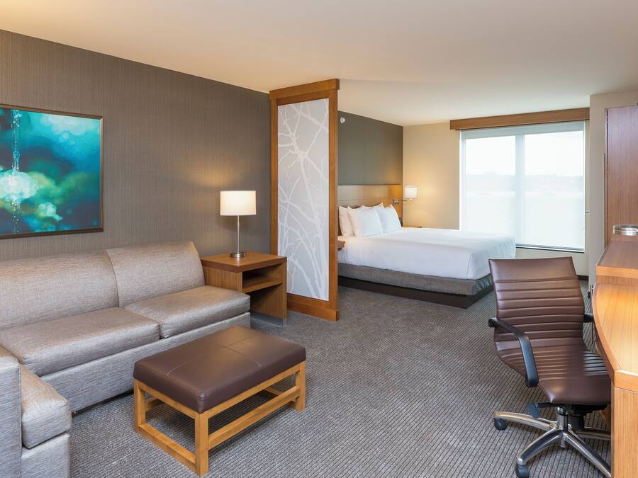 Ein Hotelzimmer mit einer Couch, einem kleinen Holztisch, einem Schreibtisch mit Stuhl, einem Bett, einem Fernseher und einem großen Fenster.