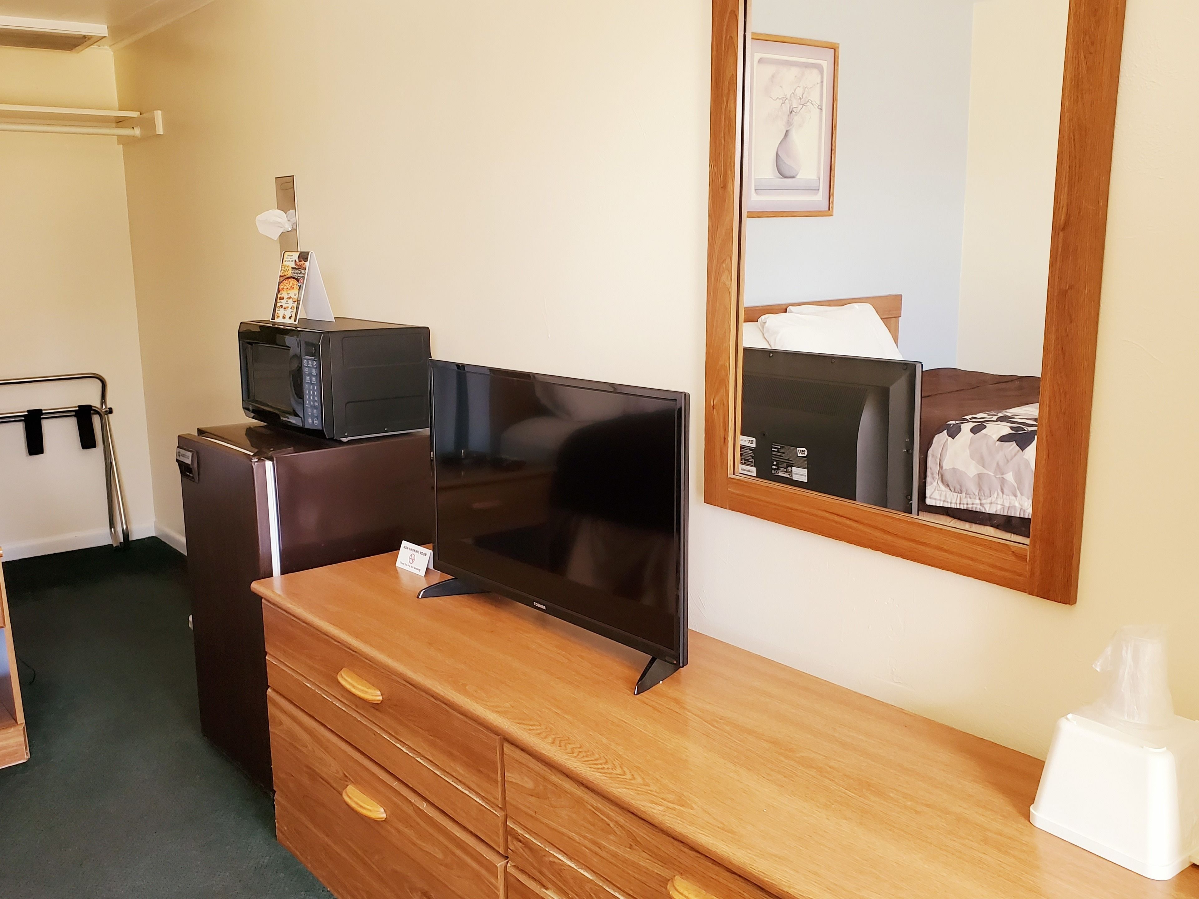 A hotel room with a wooden dresser, a flat-screen TV, a microwave, a small refrigerator, a bedside table with a tissue box, a framed picture on the wall, and a bed with a patterned bedspread.