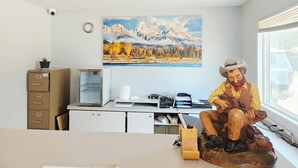 A room with a desk, a mounted painting of a mountain landscape, a wooden sculpture of a seated figure, and a window with a view of a parked car. - Budget Inn Express (Sheridan)