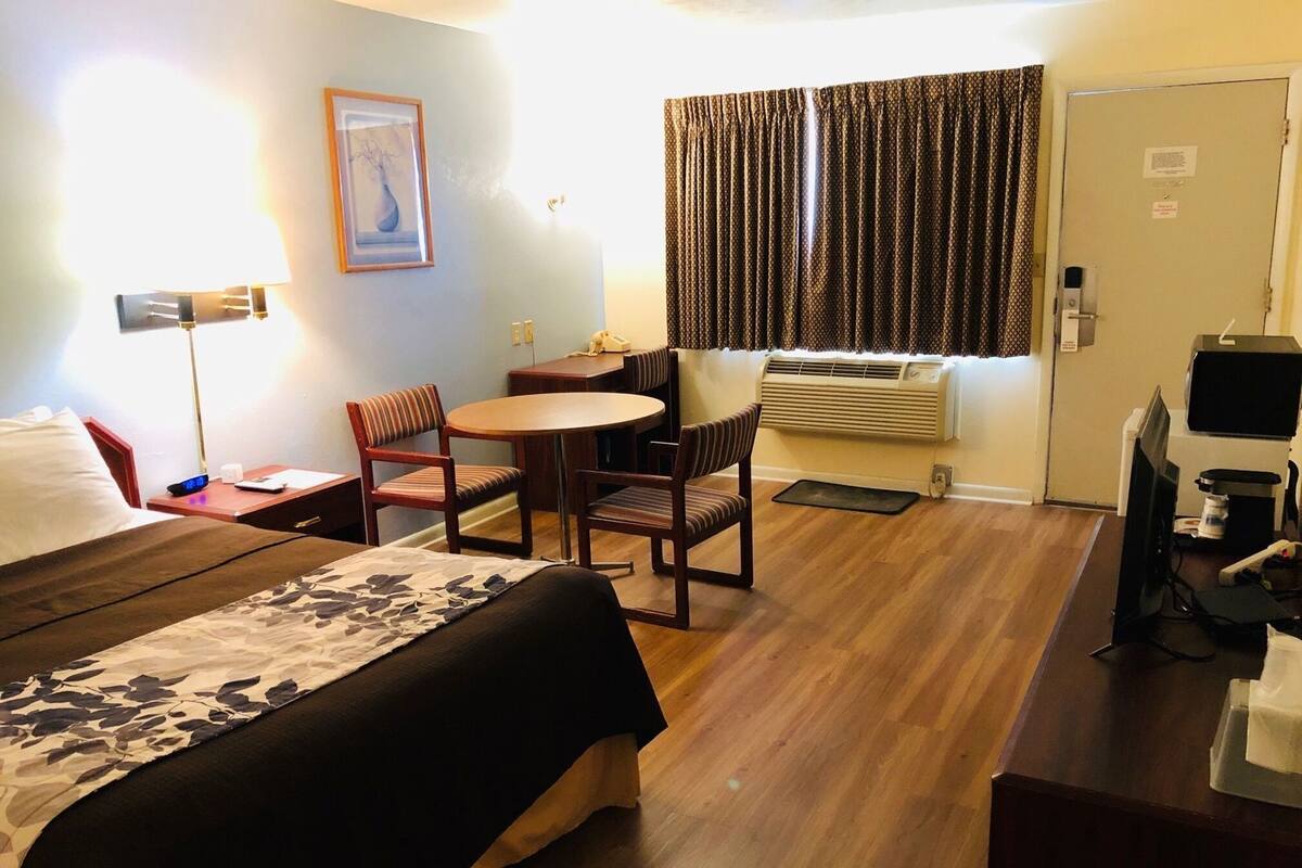 A hotel room with a bed, a desk, a chair, a small table, a television, and an air conditioning unit.